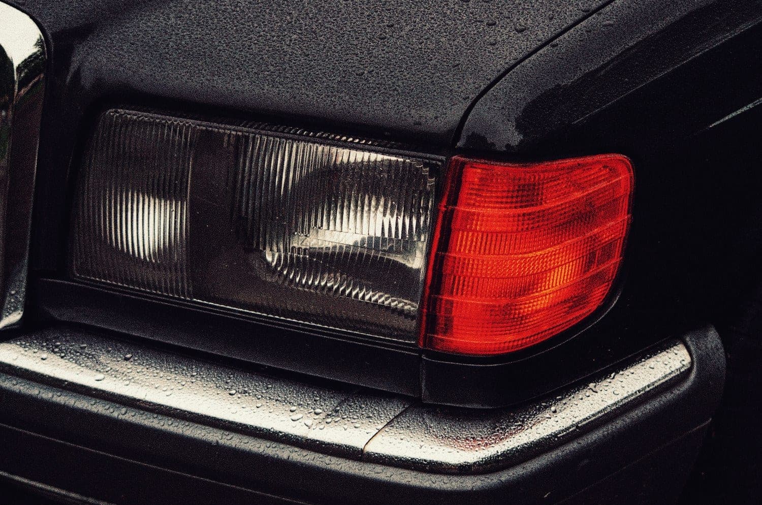 Rain-beaded tail light and trunk corner of an older black sedan