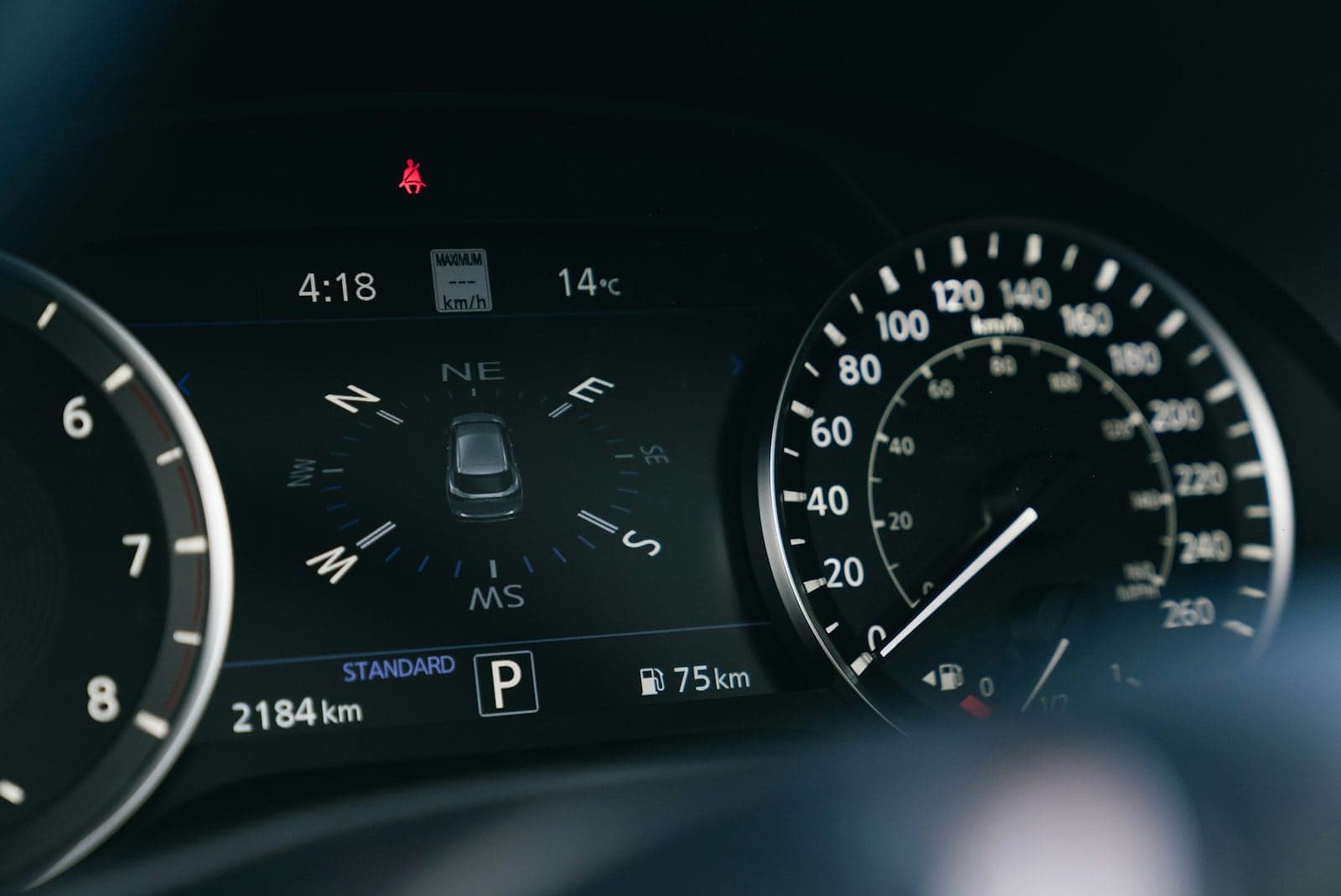 Car instrument cluster at night showing the speedometer, a digital trip display and a red seat belt warning light